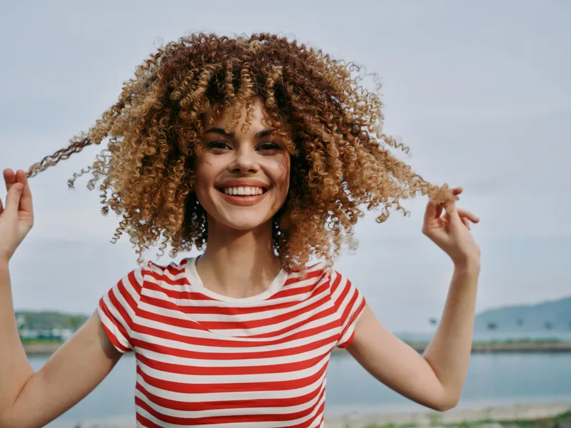 A joyful young woman with curly hair wearing a striped red shirt stands outdoors by a calm shoreline, radiating energy, confidence, and natural charm.