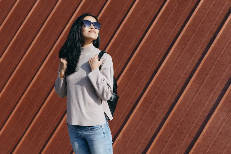 Woman wearing casual sweater and jeans with backpack and sunglasses against a diagonally planked wall, street fashion with confident pose, outdoor lifestyle image, modern urban style