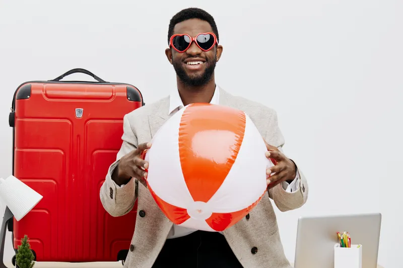 Happy cheerful man with sunglasses holding beach ball and red suitcase in studio image. Casual summer vacation concept, travel and leisure mood, bright natural lighting, fashion style, modern lifestyl