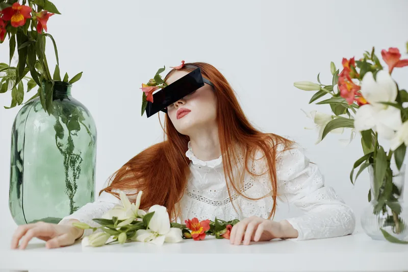 Young woman with red hair in white dress, wearing black sunglasses, surrounded by flowers, evoking a sense of calm and elegance in a minimalistic setting