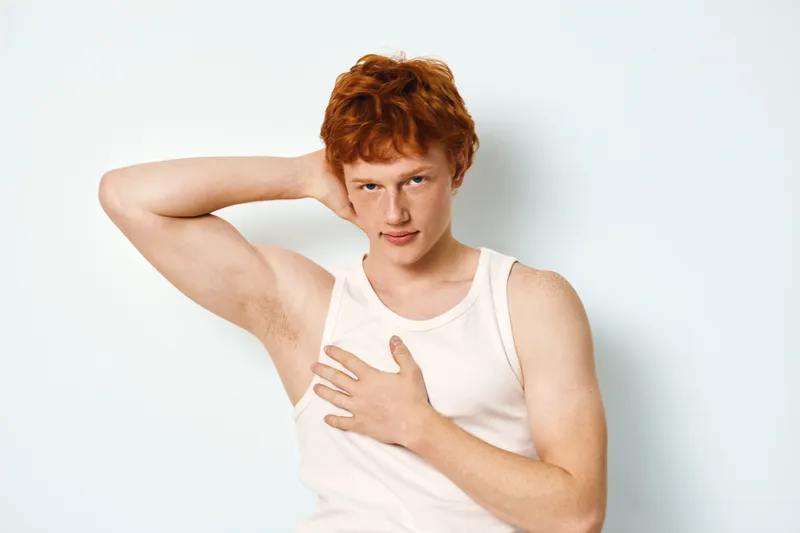 Young man with red hair in white tank top posing with hand behind neck and other on chest against white background. Casual style and confident expression portrait.
