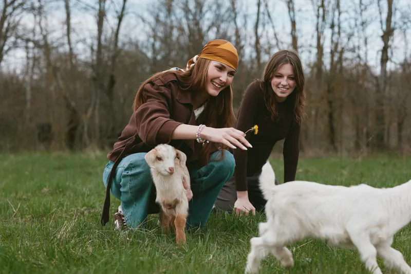 Two girl friends enjoy a sunny outdoor moment with a goat kid in a grassy field, sharing a warm, casual lifestyle as the animal wanders the meadow nearby, creating a happy, candid scene.