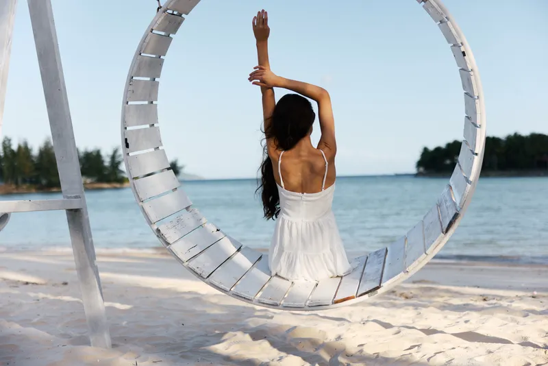 Woman in white dress sitting on a swing by the beach, relaxing under clear skies with calm water and tropical trees in the background.
