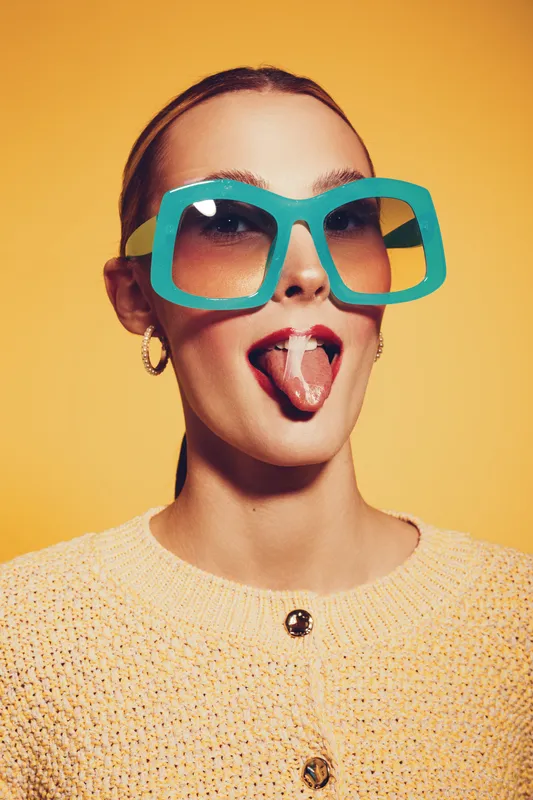 Young woman with oversized turquoise sunglasses and beige sweater sticks out tongue against yellow background. Concept of playful attitude, fashion, and summer style portrait.
