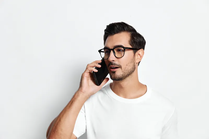 Handsome young man in glasses talking on mobile phone while standing against a plain white background. Modern lifestyle and communication concept.