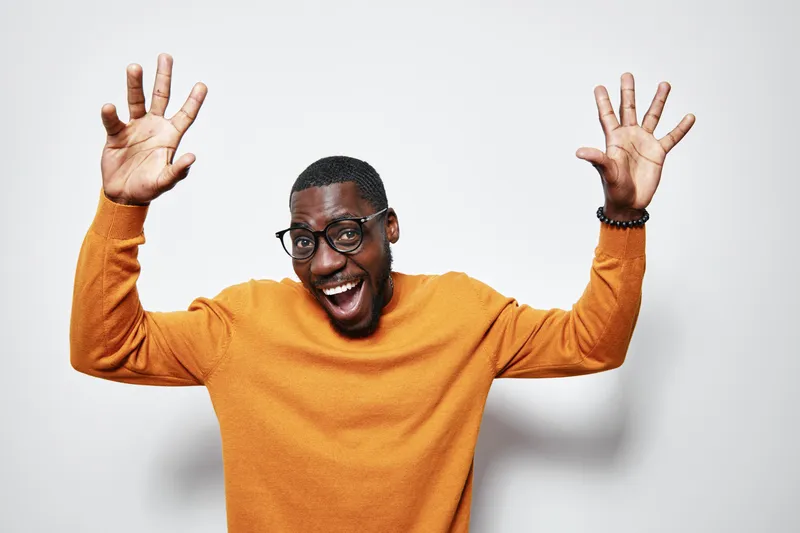 African American man with dark skin wearing glasses and orange sweater smiling and raising hands in excitement on white background. Happy male portrait with joyful expression.