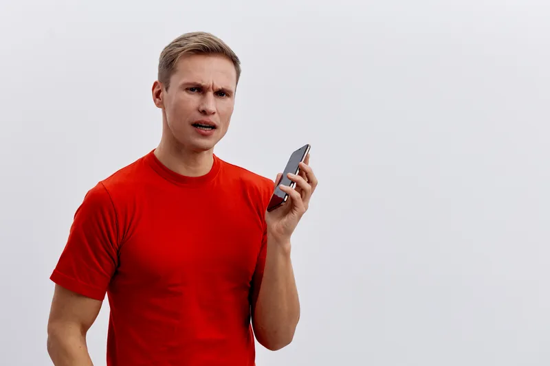 frustrated man in red t-shirt holding smartphone talking concerned about cybersecurity threat on plain white background
