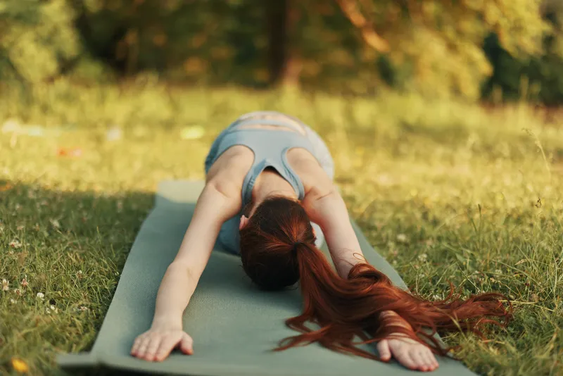 Young woman practicing yoga outdoors in nature, demonstrating relaxation and mindfulness on a green mat surrounded by lush greenery and sunlight