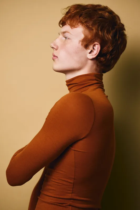 Redhead man in brown turtleneck with crossed arms posing in profile against beige background. Young adult male, side view, casual style and natural hair color with serious expression.