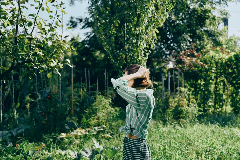 A person stands in a lush garden, arms raised to touch hair, wearing a striped top and light dress, bathed in warm sunlight.