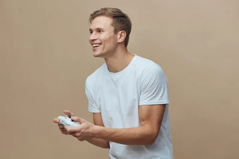 Smiling young man with casual white t-shirt playing video game controller, enjoying leisure time, indoor setting, friendly mood, lifestyle concept, happiness, technology, relaxed pose.