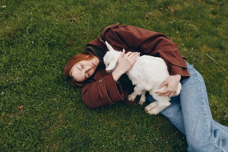 Cheerful woman lying on grass outdoors, embracing a white goat kid, sunlit moment, relaxed posture, gentle breeze, natural colors, tender affection in a pastoral setting