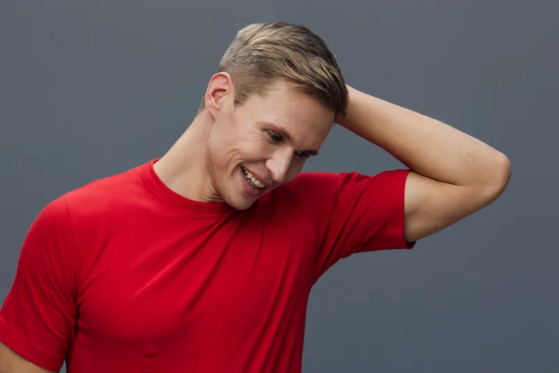 Smiling man with short hair wearing casual red t-shirt, looking down and laughing while touching his head, isolated on plain gray background, studio portrait, people lifestyle concept, cheerful and re