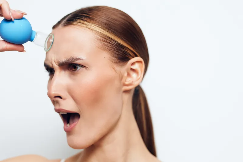 Surprised woman with a blue bulb syringe pressed to her forehead, dramatic facial expression, open mouth, studio shot on a white background, bold emotion and playful humor