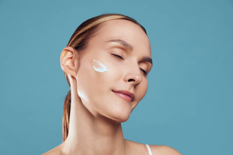 A calm young woman with blonde hair, her eyes closed, applies cream to her cheek against a blue backdrop, illustrating a skincare moment and self care.