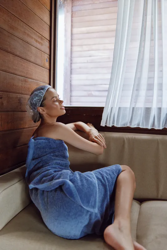 Relaxed woman wrapped in blue towel sitting near window in wooden sauna, wearing shower cap and enjoying serene calm atmosphere with soft natural light illuminating her face.