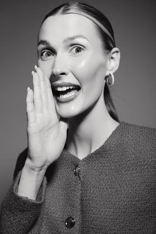 Young white woman with fair skin and blonde hair whispers with hand near mouth, wearing knitted sweater. Concept for communication and secret sharing.