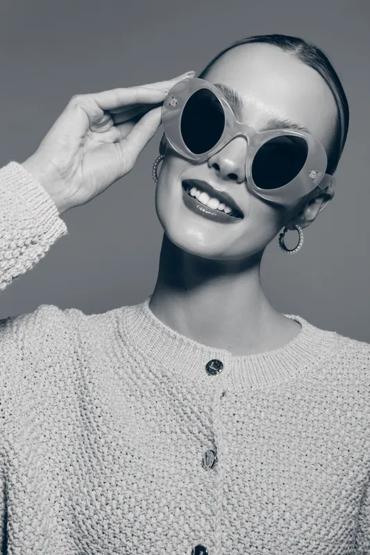 Smiling Caucasian woman with light skin wearing oversized sunglasses and knitted sweater posing in studio. Concept for fashion, accessories, and cheerful lifestyle portrait.