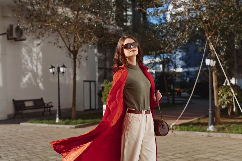A confident woman wearing sunglasses and a flowing red coat walks outdoors in a sunny urban setting with autumn trees and modern buildings in the background.