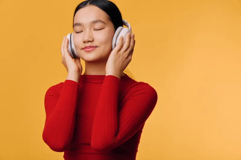 Young woman with closed eyes enjoying music wearing headphones and red sweater on orange background. Relaxed person feeling calm and happy while listening to audio.
