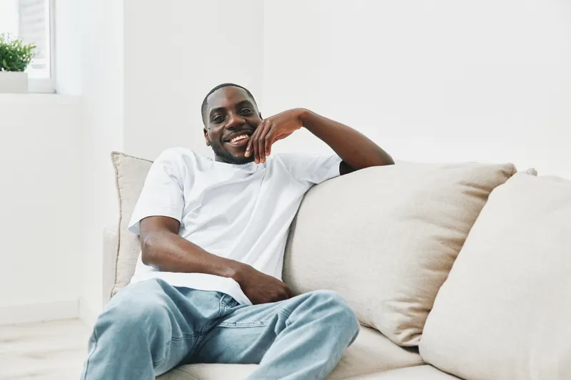 Smiling African man relaxing on a beige sofa, dressed in a casual white t shirt and blue jeans, exuding happiness in a bright, minimalist living room