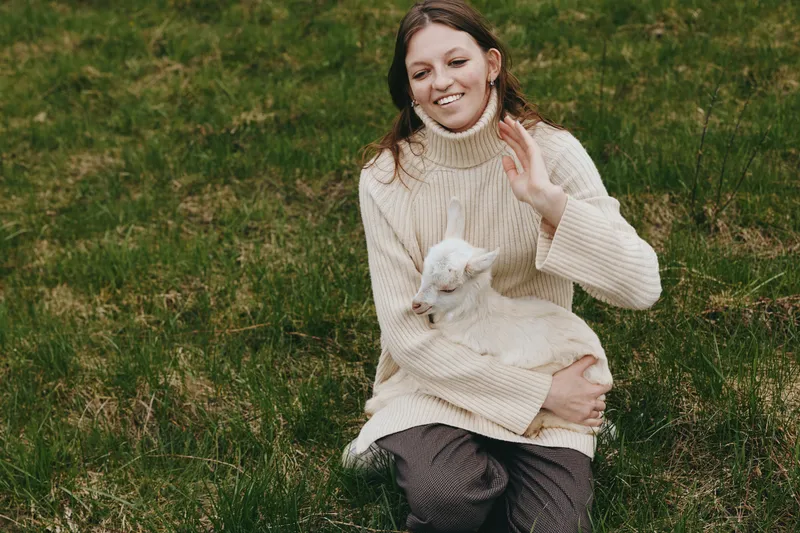 Young woman in a cozy cream sweater sits on grass, holding a goat kid. Outdoor lifestyle moment captures warmth, care, and a gentle bond with nature in a bright, peaceful setting.