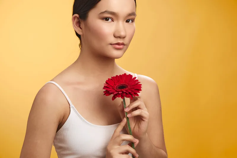 asian woman in a white tank top holds a vibrant red flower against a warm yellow backdrop, presenting a calm, intimate portrait with soft lighting and a subtle smile.