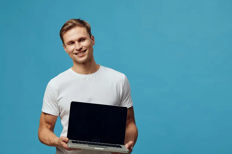 Young smiling man with light brown hair wearing white t-shirt hold open black laptop computer in hands, posing against plain blue background, studio shot, casual style, technology, communication, happ