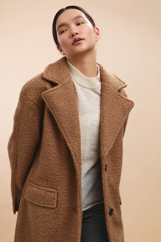 Asian woman in a warm coat stands in a neutral studio setting, exuding calm confidence and style, inviting viewer interest with a soft, modern fashion vibe.