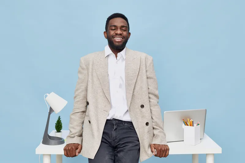 Confident young African American man standing behind a modern office desk, smiling and looking directly at camera, wearing smart casual outfit with blazer, white shirt, and jeans, isolated on solid li