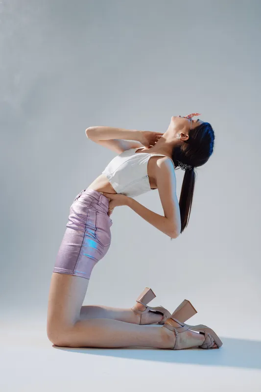 Young woman in a white top and shiny pink skirt poses on her knees with head tilted back. Studio shot on a light background. Concept of beauty, fashion, and elegance.