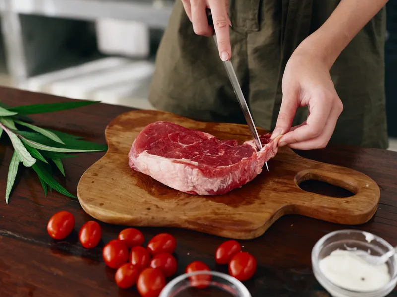 Женщина с ножом в руке нарезает свежее стейковое мясо для жарки на кухне