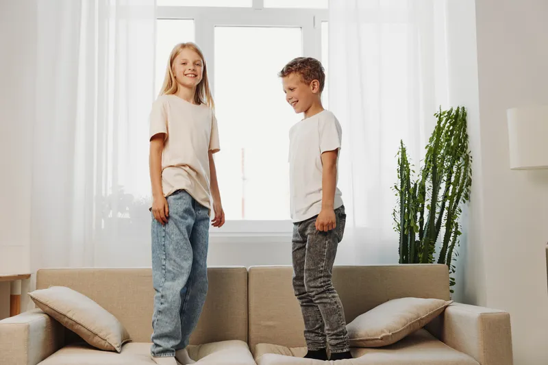 Happy children jumping on sofa in bright living room, smiling boy and girl enjoy playful moment together at home, casual clothes and natural light.