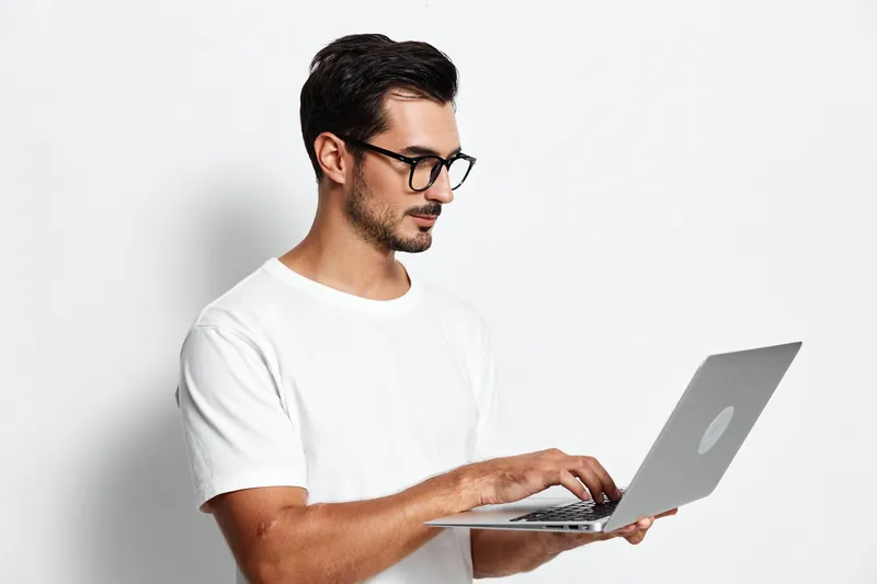 Focused young smiling man wearing glasses working on laptop, standing against plain white background. Professional lifestyle and productivity concept.