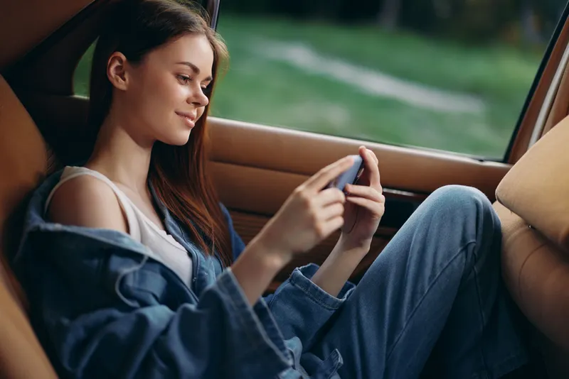 Young woman relaxing in a car, wearing casual denim clothes, absorbed in her phone, showcasing a serene expression against a natural green background.