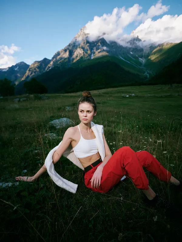 Woman resting on a meadow with mountains in the distance, wearing a white top and red pants, scarf over her shoulder, showcasing casual outdoor fashion and natural scenery