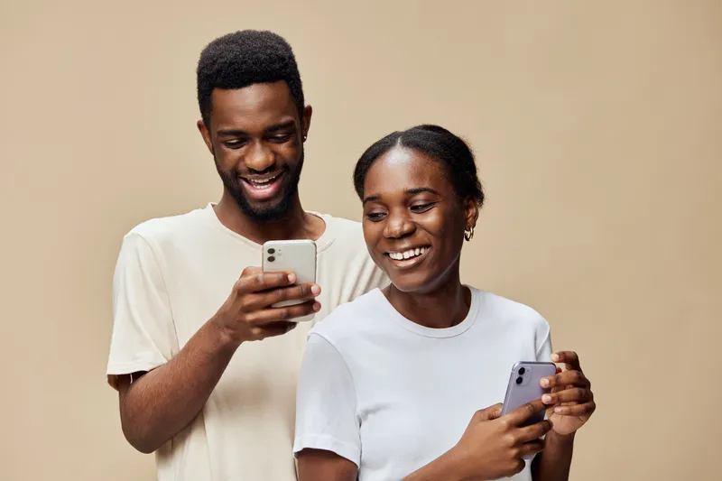 Young African couple smiling while using smartphones together against a beige background, showcasing modern technology and joyful emotions