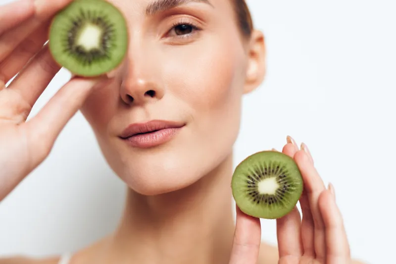Young woman with clear skin holds two halves of fresh kiwi fruit near her face against a white background, expressing natural beauty and healthy lifestyle with a calm and confident expression.