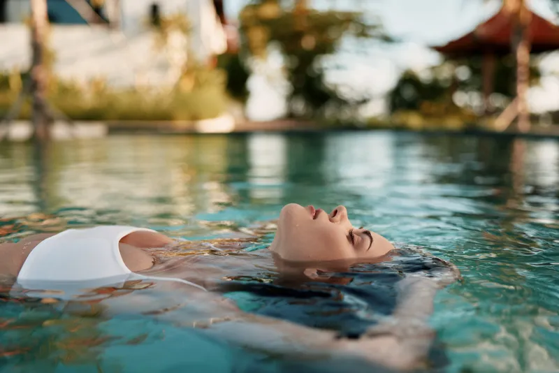 Relaxed woman floating in a sunlit pool, eyes closed, peaceful smile, white swimsuit, gentle breeze, clear water, vibrant surroundings, carefree lifestyle, soft reflections, natural colors
