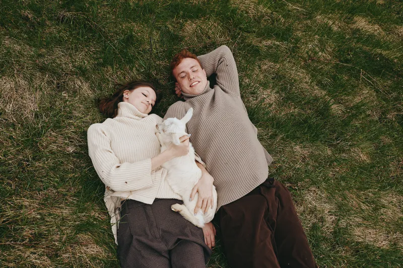 Cheerful couple outdoors on grass with a goat kid, relaxing, lying close, wearing sweaters, neutral tones, cozy mood, sunlit countryside scene in a peaceful field with soft breeze and colors