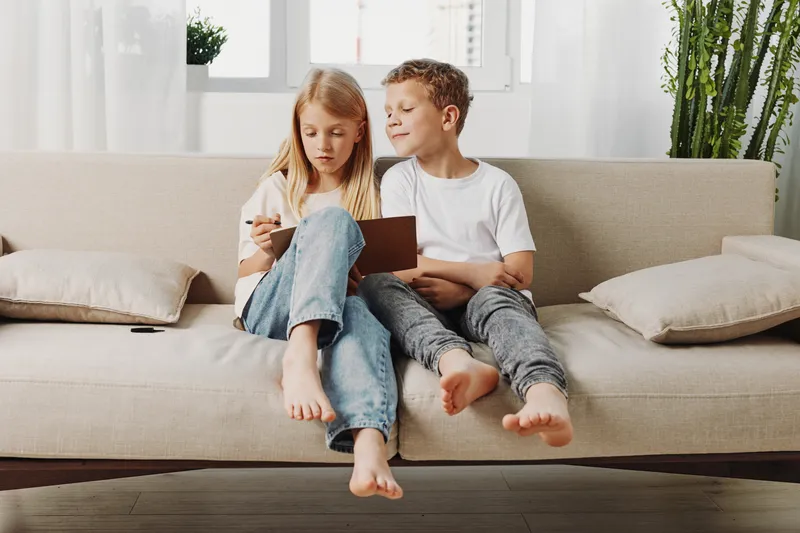 Two barefoot children sitting on a beige sofa playing with a tablet in a bright living room with plants and cushions.