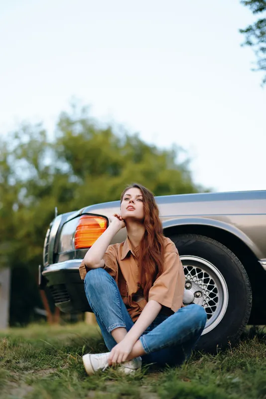 Young woman sitting near vintage car in casual outfit, showcasing relaxed style and thoughtful expression amidst outdoor nature.