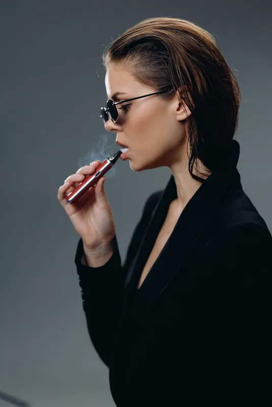 Stylish woman vaping with e-cigarette in black outfit and sunglasses, modern and edgy look with dark background.