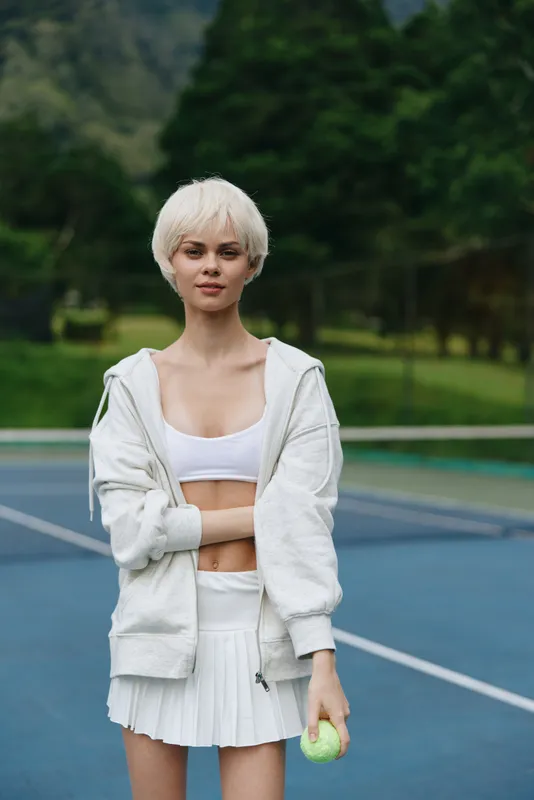 Young female tennis player with short white hair in white sportswear, exuding confidence while holding a tennis ball on a court surrounded by greenery