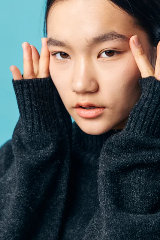 Young woman with clear skin and expressive eyes posing with hands near face wearing dark sweater on blue background. Beauty, skincare, fashion and portrait concept.