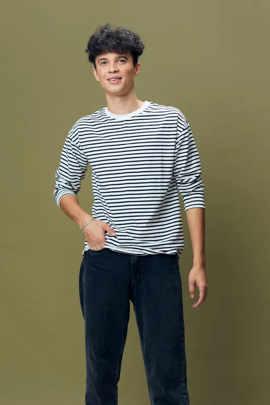 Young man wearing striped long-sleeve shirt and black pants standing against olive green background with relaxed and friendly expression