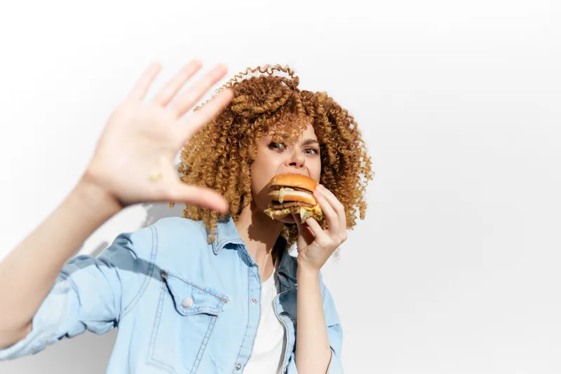 Молодая женщина с вьющимися волосами держит бургер близко к лицу, но при этом отвергает его