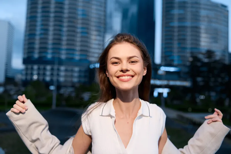 Smiling woman in white blouse enjoying urban evening with modern buildings background, positive emotion, casual style, natural light, city lifestyle concept