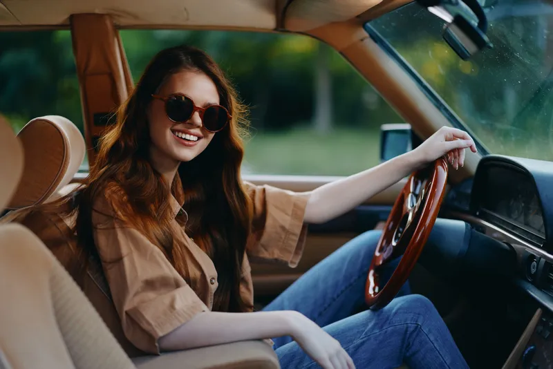 Cheerful young woman driving a vintage car, wearing stylish sunglasses and a casual outfit in a lush green outdoor setting.