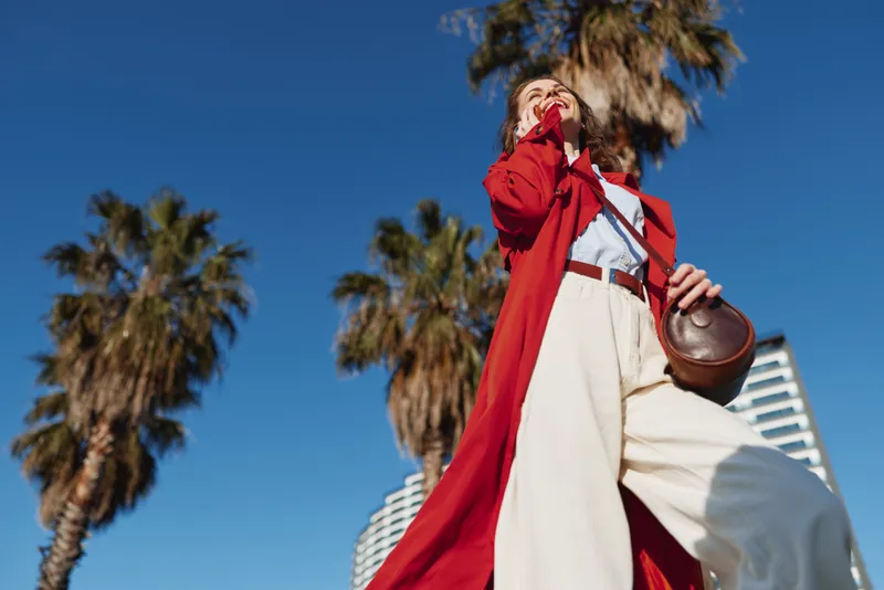 A stylish woman in a red trench coat and white blouse stands outdoors beneath palm trees, looking up with a confident pose.